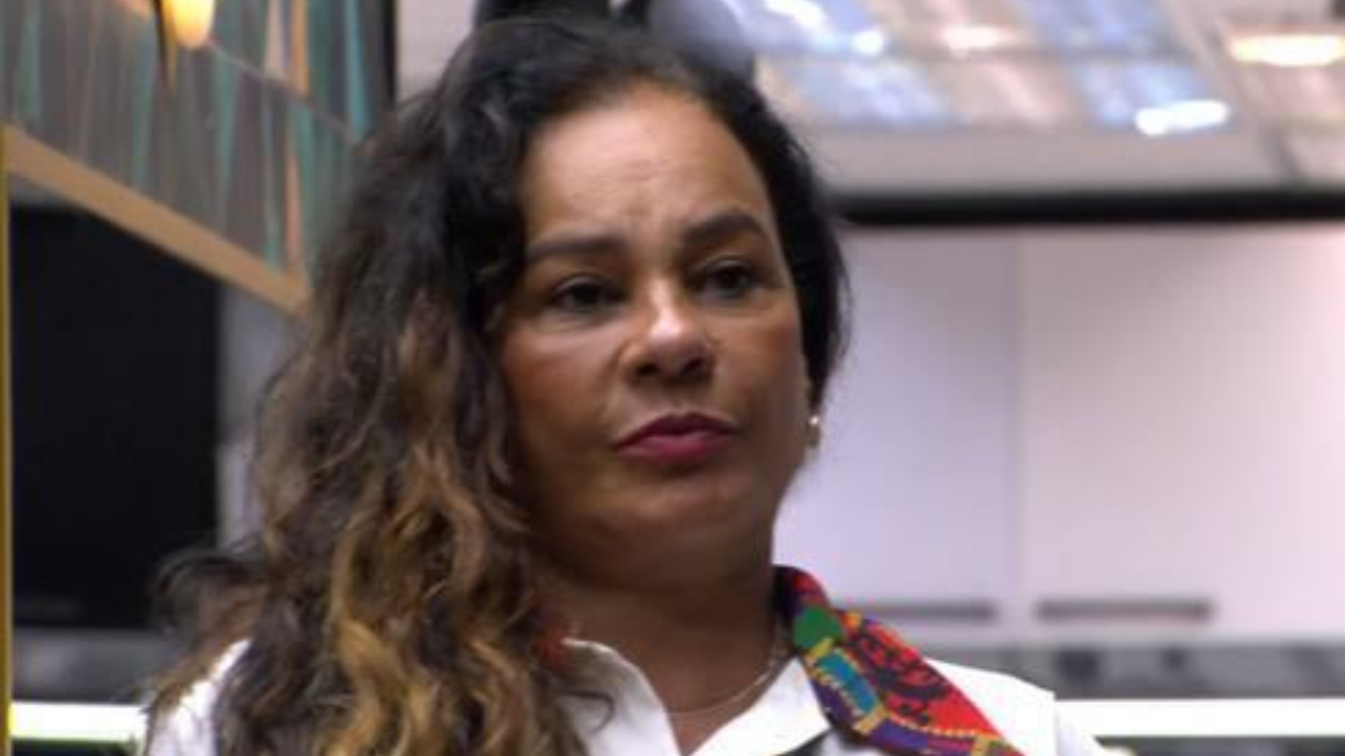 Mulher de cabelos cacheados e escuros, usa um colar e um lenço colorido no pescoço. Ela está em um ambiente interno com ilum…