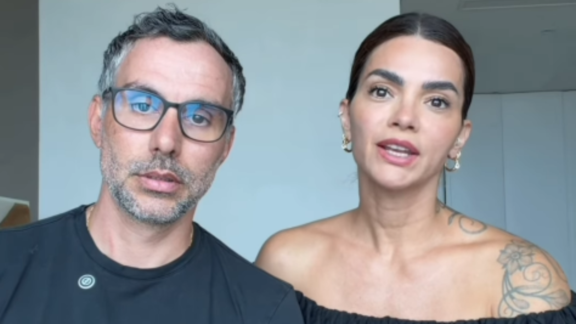 Casal formado por homem de óculos e mulher com tatuagens no ombro, olhando para a câmera em um ambiente interno.