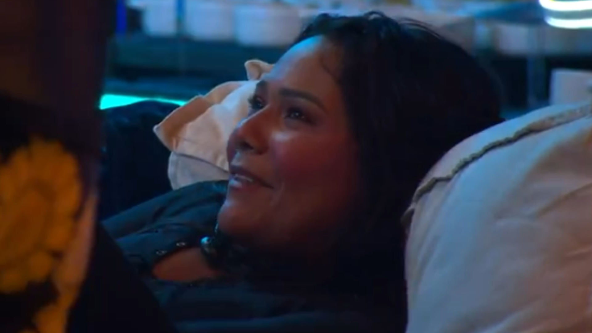 Mulher deitada em uma cama, com maquiagem e cabelo escuro, olhando para o lado com um sorriso.